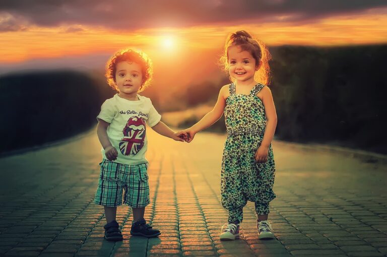 Two young children holding hands and smiling while walking on a sunlit path at sunset.
