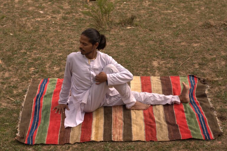 Person in white clothing performing a seated yoga stretch outdoors on a striped mat.
