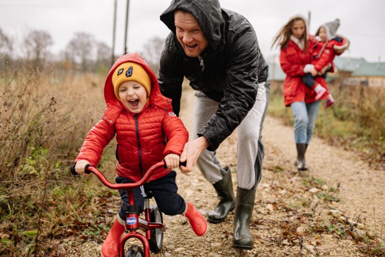 Child in a red jacket riding a small bike with adults walking beside.
